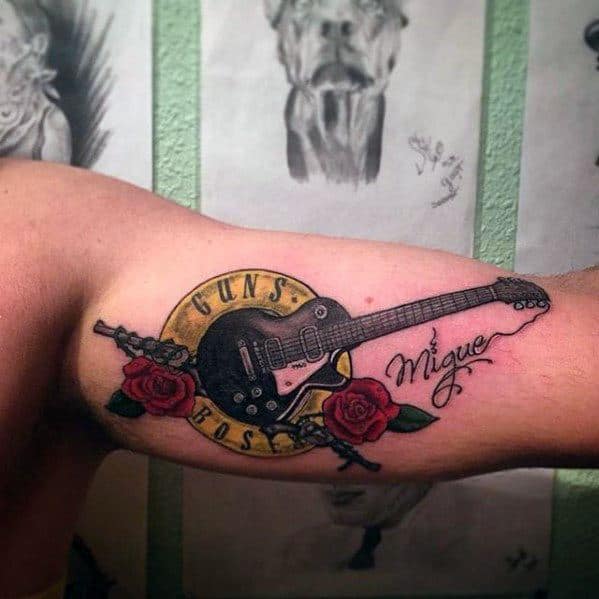 Tattoo of a black electric guitar over a yellow "Guns N' Roses" circle, with red roses, bullets, and the name Miguel on the bicep.