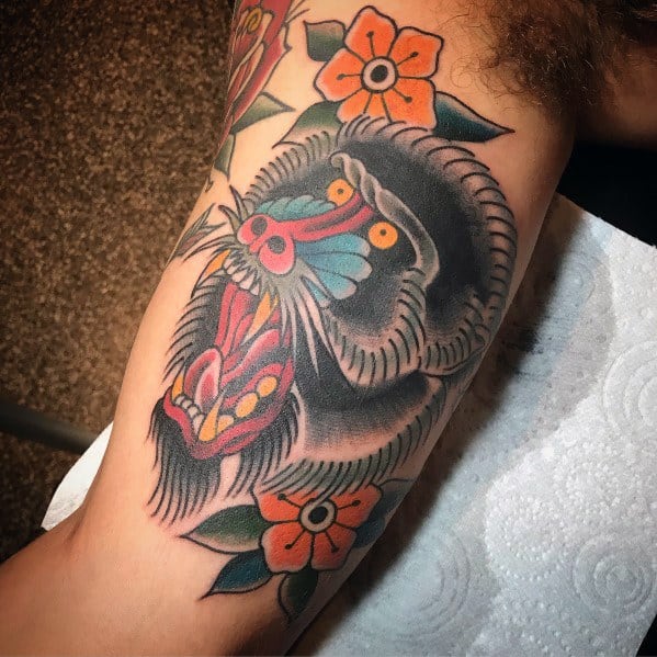 Neo-traditional baboon tattoo with bold outlines, vivid colors, and fierce expression, accented with floral elements for a striking arm design.