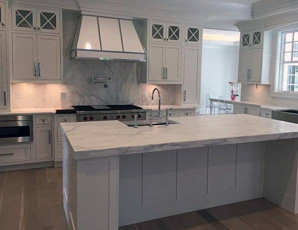Light grey kitchen with marble island, stainless steel range hood, and bright under-cabinet lighting.