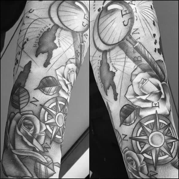 Forearm tattoo with a magnifying glass over a nautical map, compass, roses, and leaves, creating a detailed black and gray design.
