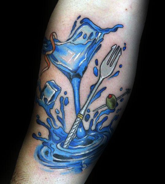 Blue martini glass tattoo with a splash effect, featuring a fork, olive, and ice cubes. A bold and dynamic design on the forearm.