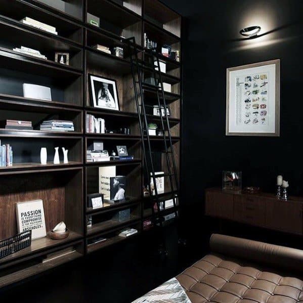 Dark-toned home library with a ladder, wooden shelves, books, framed art, and a brown leather chair