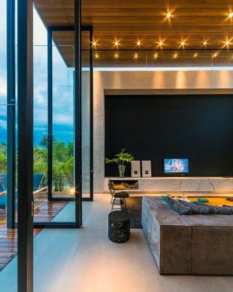 Modern living room with large glass walls, sleek furnishings, recessed lighting, and visible outdoor greenery