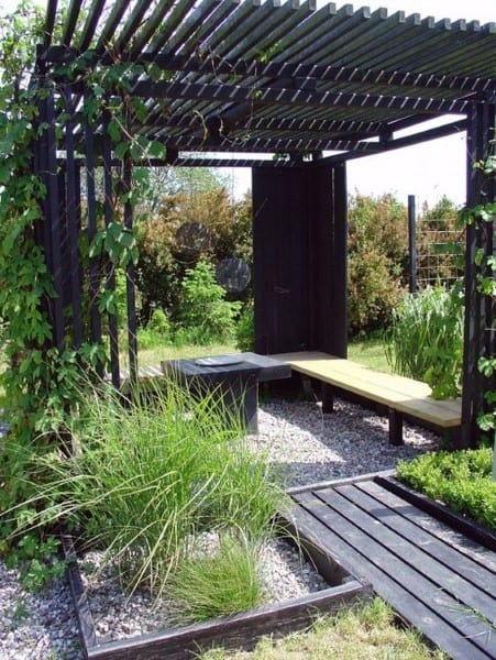Rustic pergola with slatted roof, wooden bench, gravel flooring, and lush greenery surroundings.