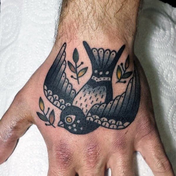 Black and grey pigeon tattoo on hand, mid-flight with leaves in its wings, done in bold traditional style with solid shading.