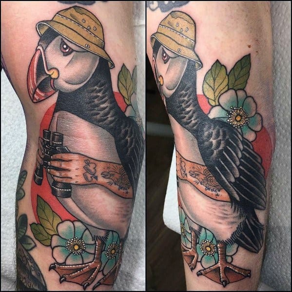 Colorful tattoo of a puffin wearing a hat and holding binoculars, surrounded by flowers and leaves, blending adventure and nature themes.