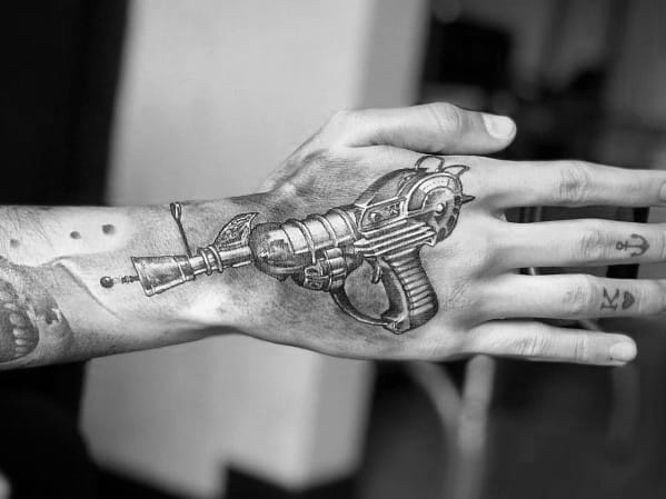 Realistic black and grey ray gun tattoo on the hand, featuring fine shading and intricate details for a bold sci-fi inspired look.