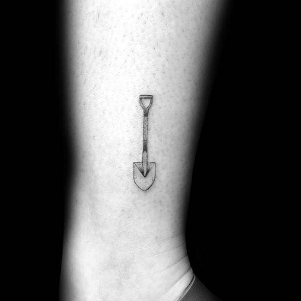 Minimalist shovel tattoo with fine black linework, placed on the lower leg, featuring a simple spade design with a straight handle.