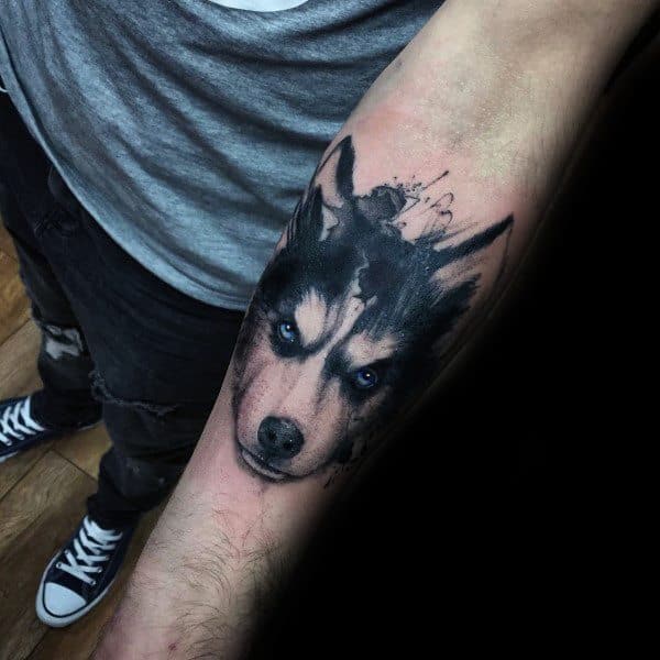 Realistic Siberian Husky tattoo on a man's forearm, featuring intense blue eyes and an artistic splash effect for a bold and striking design.