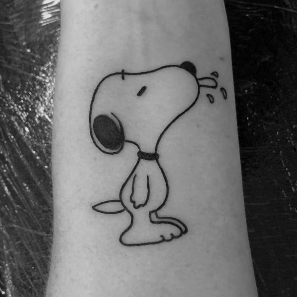 Black and white tattoo of Snoopy standing with his head tilted back, sticking out his tongue, placed on a person's forearm.