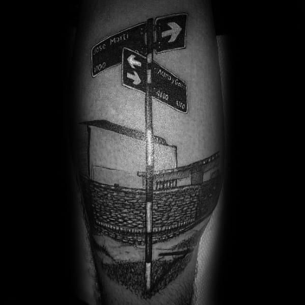 Detailed black and grey tattoo of a street signpost with arrows and street names, featuring a realistic urban background reflection.