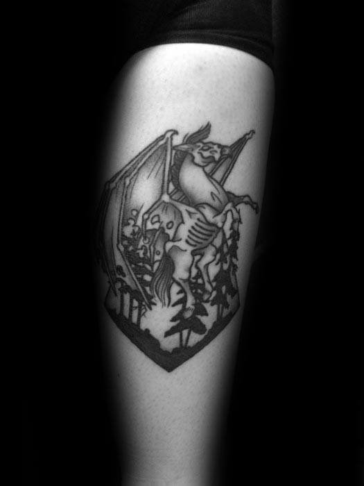 Thestral tattoo in a bold blackwork style, set within a forest scene inside a shield-shaped frame on the upper arm.