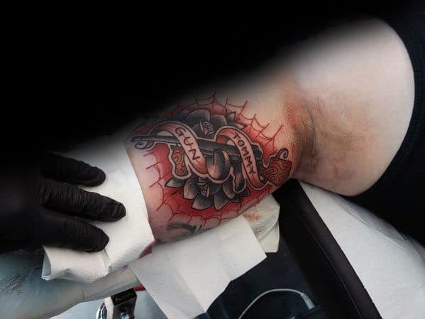 Traditional Tommy gun tattoo with bold colors and a banner wrapped around the firearm, featuring a red spiderweb background on the armpit.