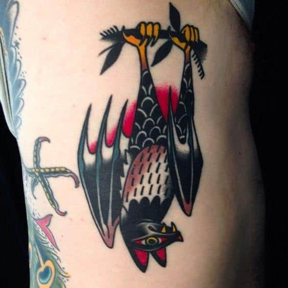 Bold traditional bat tattoo hanging upside down with red, yellow, and black ink, gripping a branch with stylized wings.