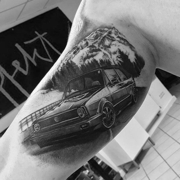 Realistic Volkswagen car tattoo in black and gray ink with a mountain landscape in the background on the upper arm.