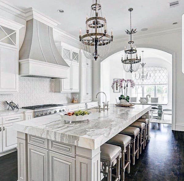 White kitchen with marble island, upholstered bar stools, and ornate chandeliers.