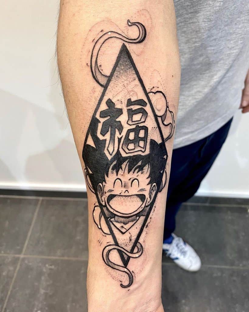 A tattoo of young Goku from Dragon Ball, smiling inside a diamond-shaped frame with the Chinese character for "fortune" (福) and a swirling cloud design.