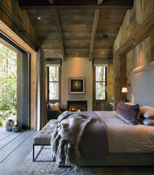 Rustic bedroom with wooden walls, fireplace, and cozy forest view.