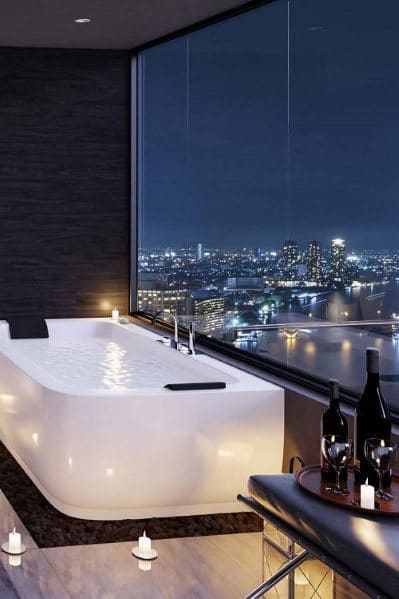 Elegant bathtub beside a window with city night view, candles, and wine
