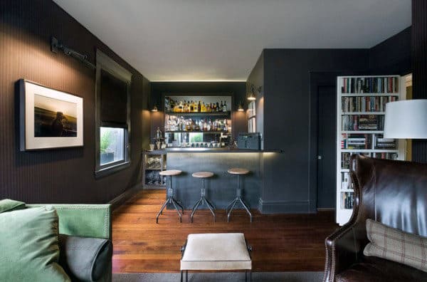 Cozy home bar with dark walls, framed art, wooden floor, three bar stools, and a bookshelf