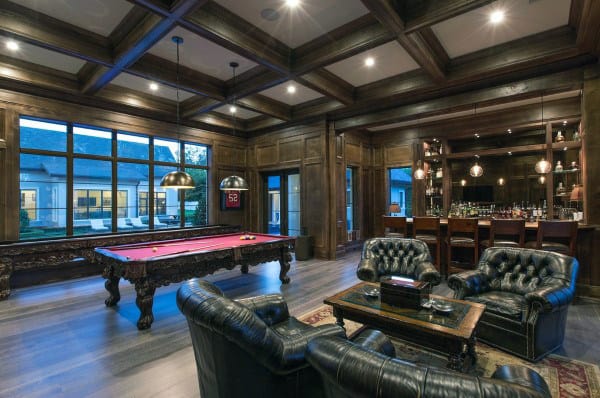 Luxurious game room with a pool table, black leather chairs, wood paneling, large windows, and a stocked bar