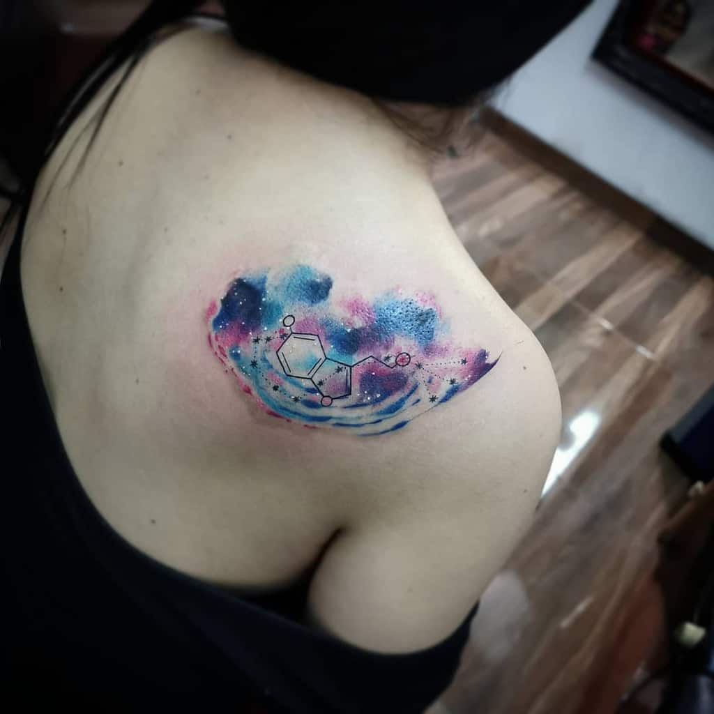 A vibrant watercolor tattoo of a serotonin chemical structure adorns the person's shoulder, blending art with scientific elegance