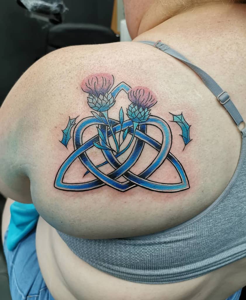 Tattoo of vibrant blue thistles intertwined with a Celtic knot elegantly graces a woman's shoulder