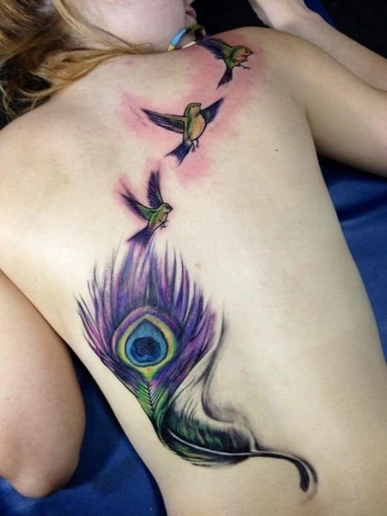 Colorful back tattoo featuring a vibrant peacock feather with shades of purple and green, transitioning into three flying birds with detailed shading.