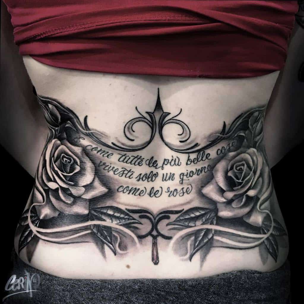 A black and white tattoo across the back featuring two detailed roses with a flowing design, accompanied by a quote in Italian that translates to "Like all the most beautiful things, you live only one day, like the roses"
