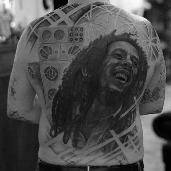 Full back tattoo of Bob Marley smiling, surrounded by speaker designs and abstract geometric elements, blending music and art in black and gray.