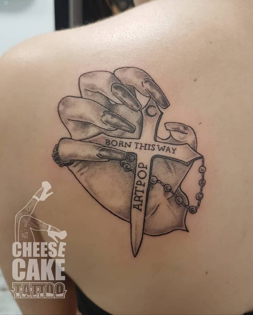 Tattoo of a hand holding a dagger with text "Born This Way" and "Artpop" on an upper back