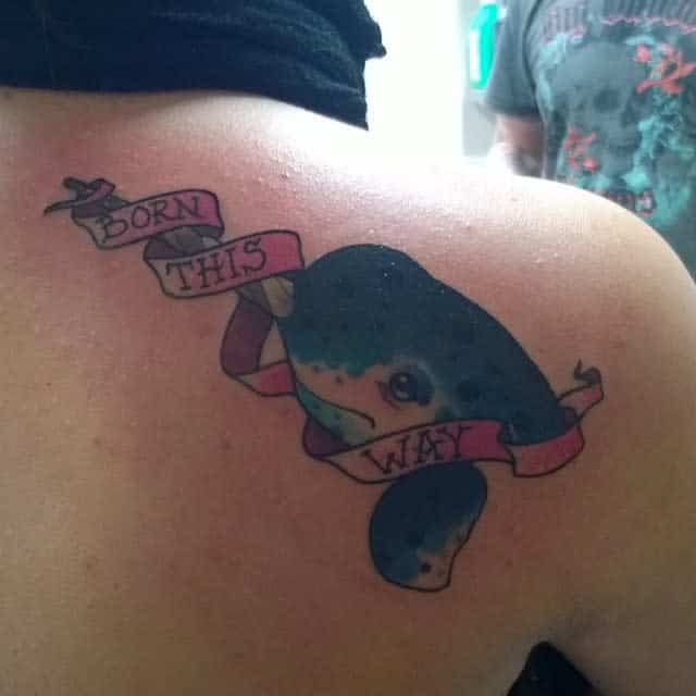 Tattoo of a whale on a shoulder with a banner reading "Born This Way" wrapped around it