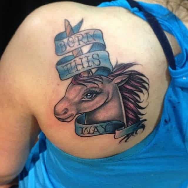 A tattoo of a unicorn with pink mane and a ribbon reading "Born This Way" on a shoulder