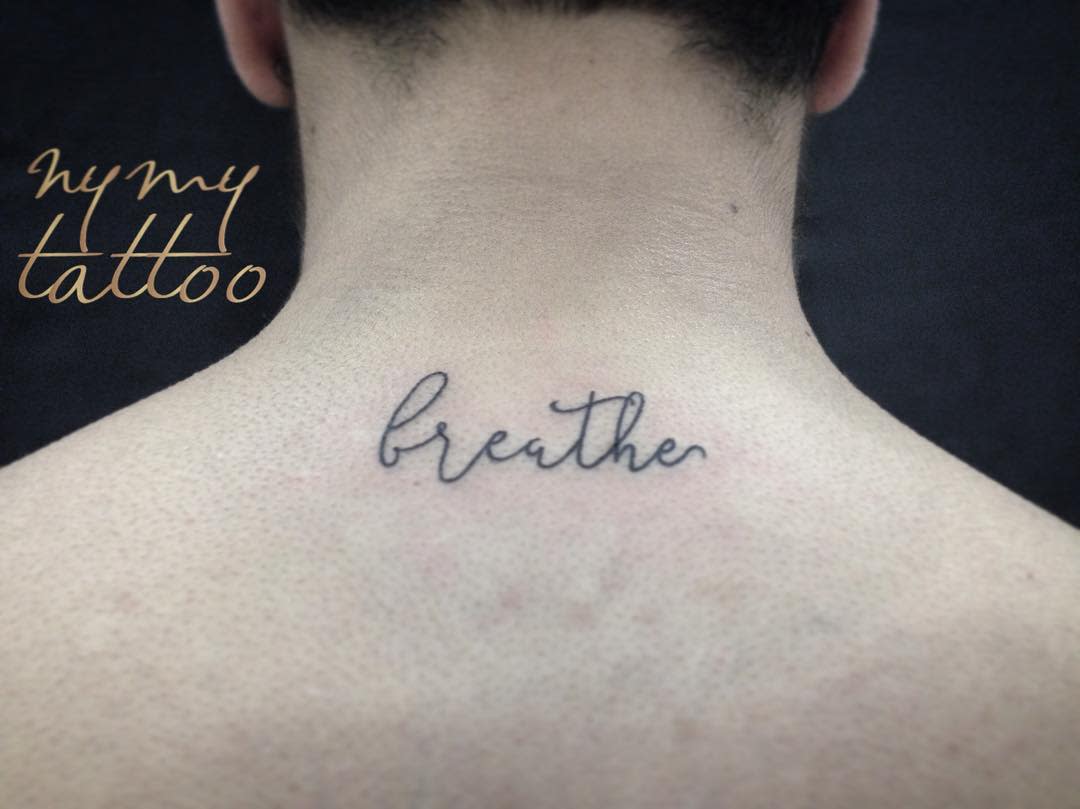 A delicate tattoo of the word "breathe" flows in elegant cursive on the back of a person's neck, serving as a gentle reminder to find calm amidst chaos