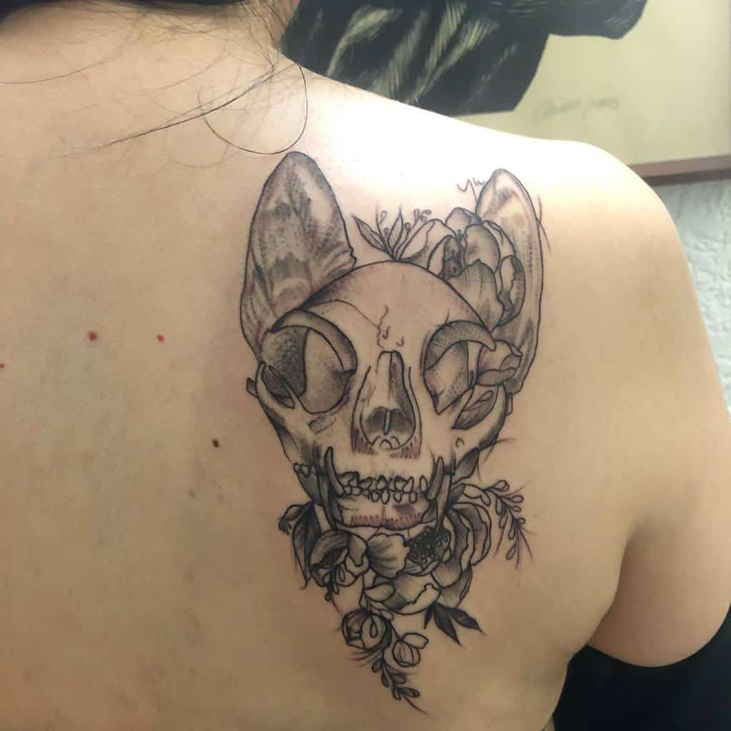 A detailed tattoo on a person's upper back features a cat skull intricately adorned with flowers