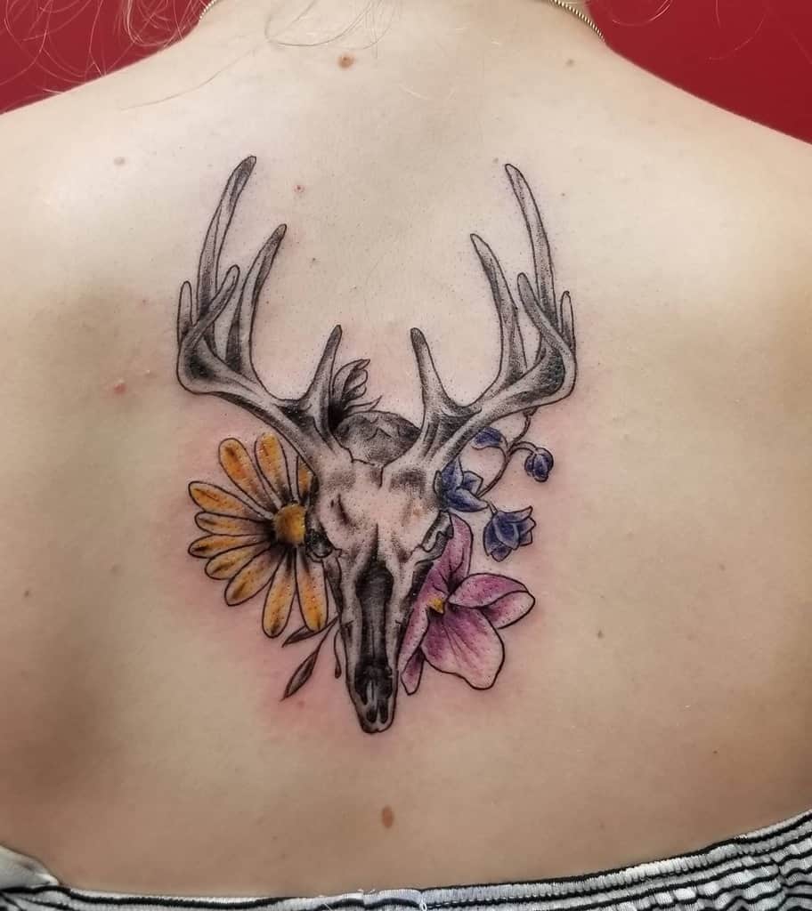 Tattoo of a striking deer skull with antlers, beautifully surrounded by delicate flowers on a person's upper back