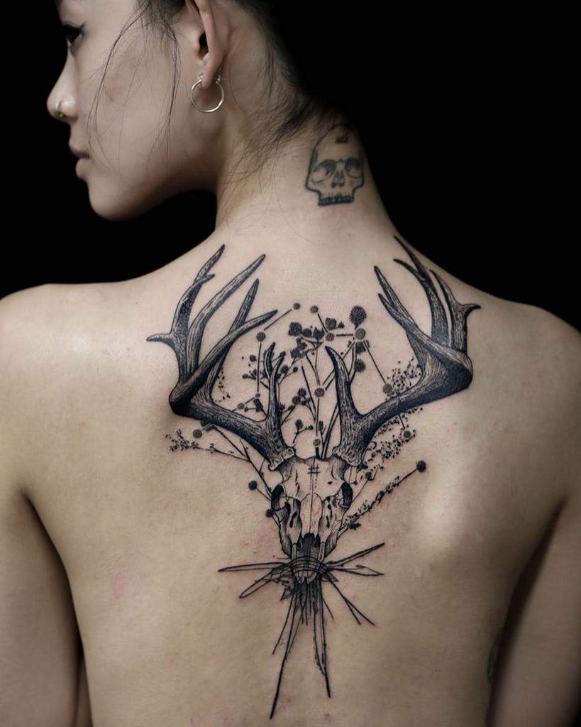 A woman with a large deer skull tattoo adorned with flowers gracing her back and a small skull etched onto her neck