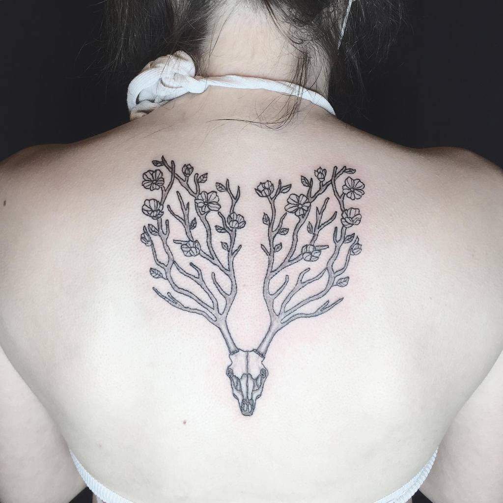 Back tattoo featuring a deer skull with antlers seamlessly transforming into blooming tree branches, creating a fusion of nature and artistry