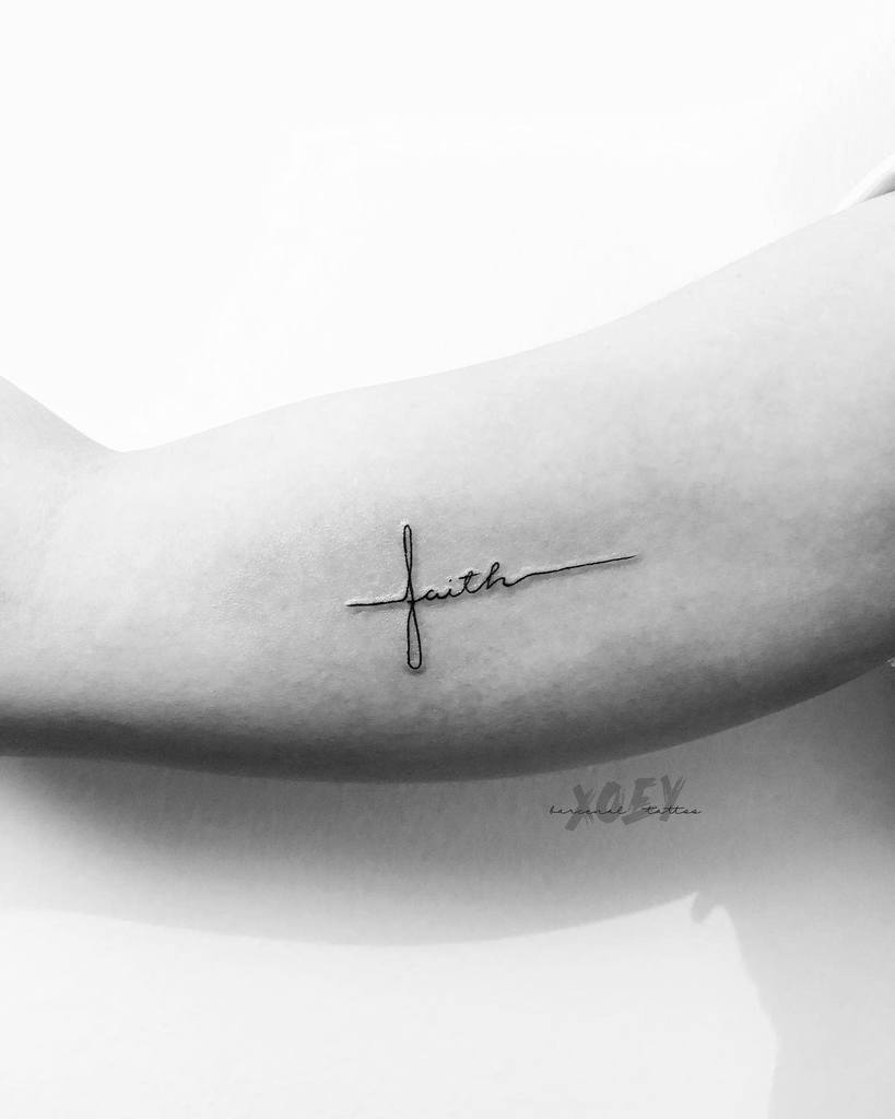 A minimalist tattoo of the word "faith" artistically entwined with a subtle cross in cursive graces an upper arm