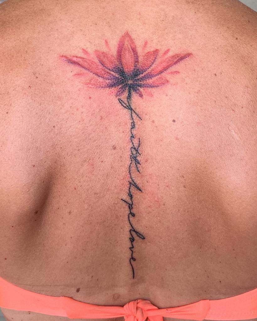 Back tattoo featuring a pink lotus flower at the top, with 'faith hope love' written in delicate cursive forming the stem of the flower, symbolizing spiritual growth and resilience