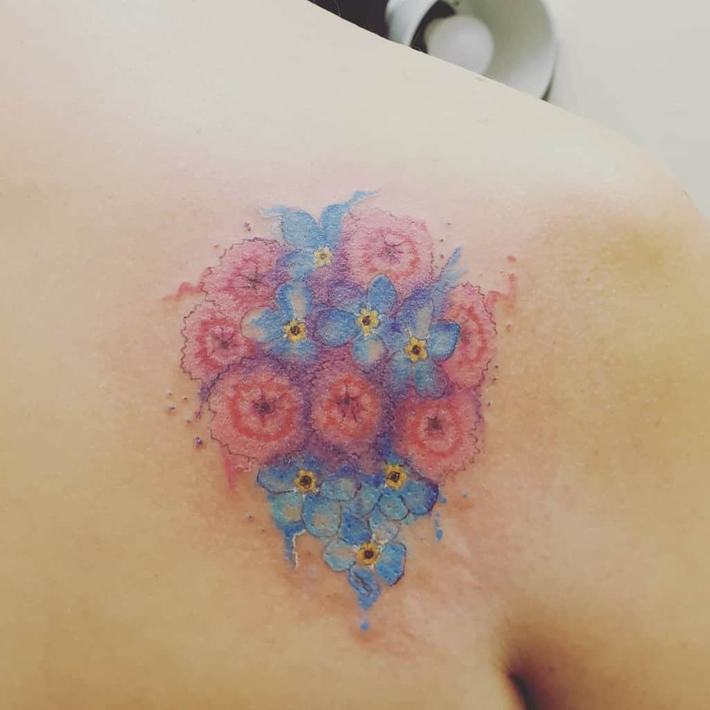 The tattoo shows a beautiful cluster of forget-me-not flowers, blending soft shades of pink and blue with watercolor effects, placed on the upper back