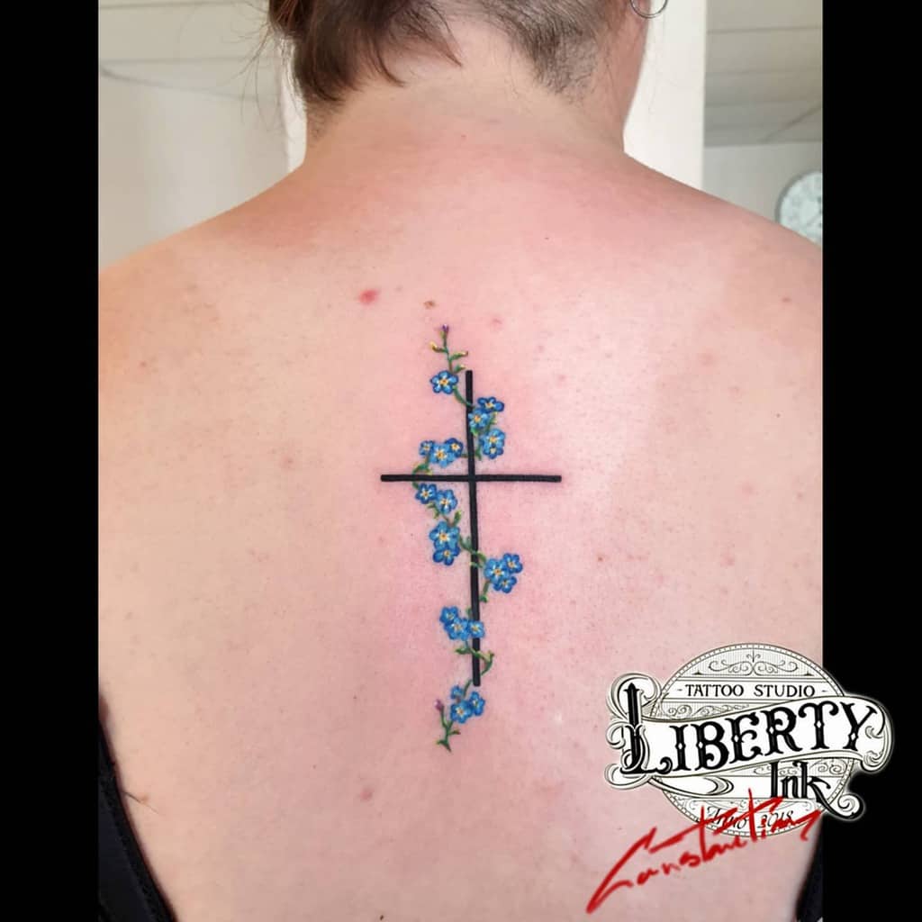 Forget-me-not flowers delicately entwine with a cross tattoo on the person's upper back, adding a touch of vibrant blue to the design