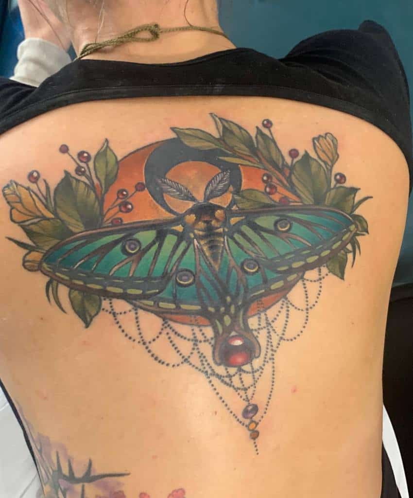A vibrant tattoo of a Luna Moth surrounded by foliage, flowers, a crescent moon, and delicate chains with jewels