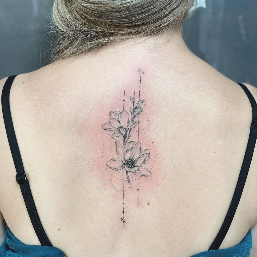 A back tattoo of delicate magnolia flowers intertwined with geometric elements graces a back