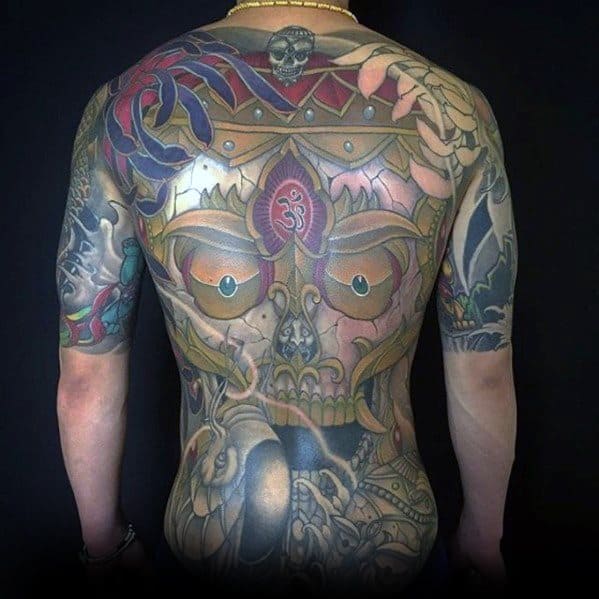 Full back Tibetan skull tattoo with fierce eyes, intricate crown, and spiritual symbols, extending to both arms in vivid detail.