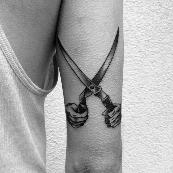 Tattoo of two detailed hands gripping large garden shears, blades crossed to form an X on the back of the arm.