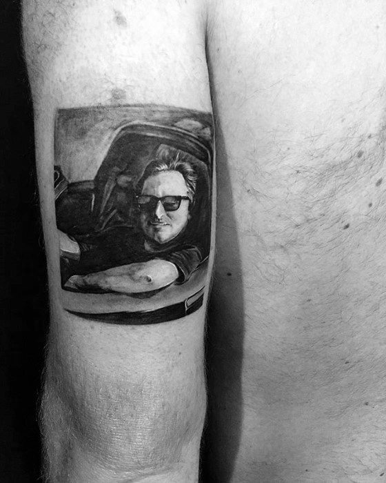 Detailed black-and-grey tattoo of a man wearing sunglasses, leaning out of a car window with a relaxed expression