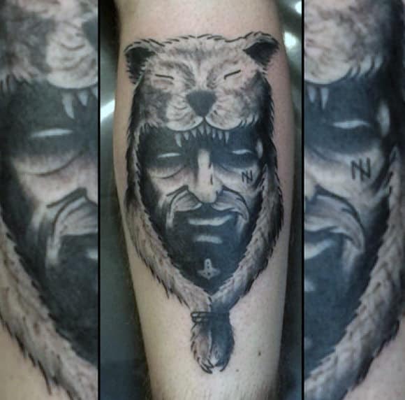 Black and grey tattoo of a warrior's face wearing a bear headdress, with intricate shading and bold details on the calf