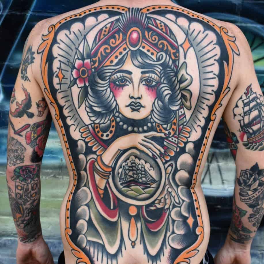 Striking full-back tattoo of a gypsy woman with an ornate headpiece, bold shading, and a crystal ball revealing a sailing ship.