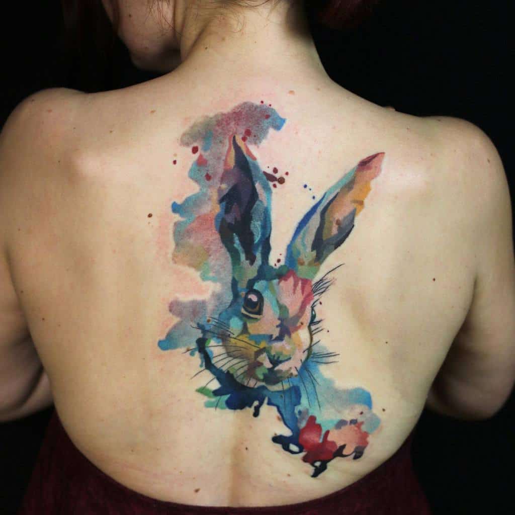 Bold watercolor tattoo of a rabbit on the back, with vibrant splashes of blue, pink, yellow, and purple creating a dynamic and artistic design